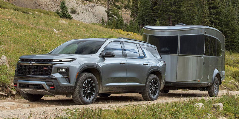 Gray Chevy Traverse 2024 towing a silver travel trailer on a dirt road in a mountainous, forested landscape.